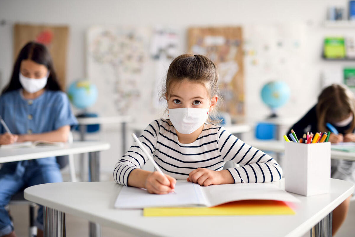 uma menina na escola, escrevendo no caderno, usando uma mascara contra o coronavirus